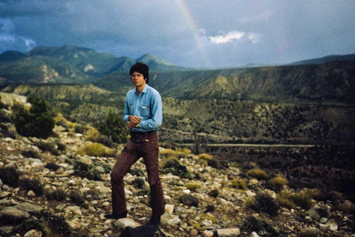 a man standing on. rocky ground with mountains and a rainbow in the background