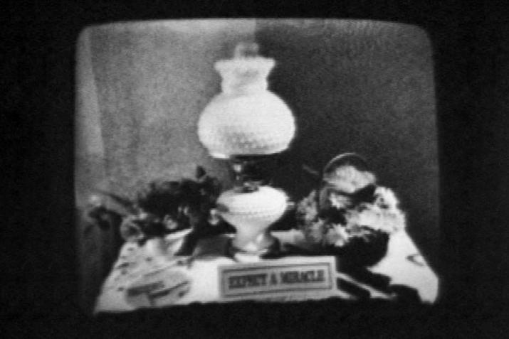 black and white image of a lamp on a table and a small sign that says "Expect a Miracle"