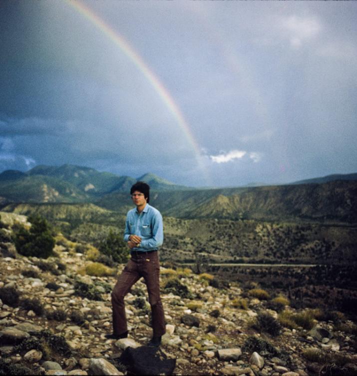 Man standing alone in foreground with rainbow and mountains in the background