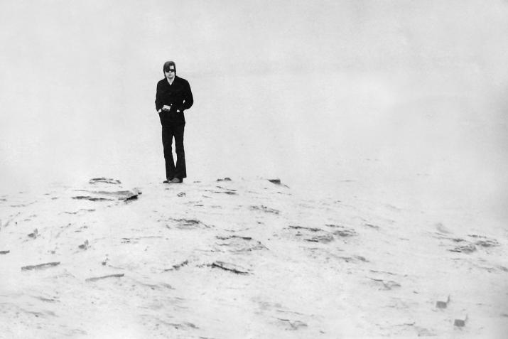 black and white image of man standing on shoreline wearing all black.  The foreground and background are very light.