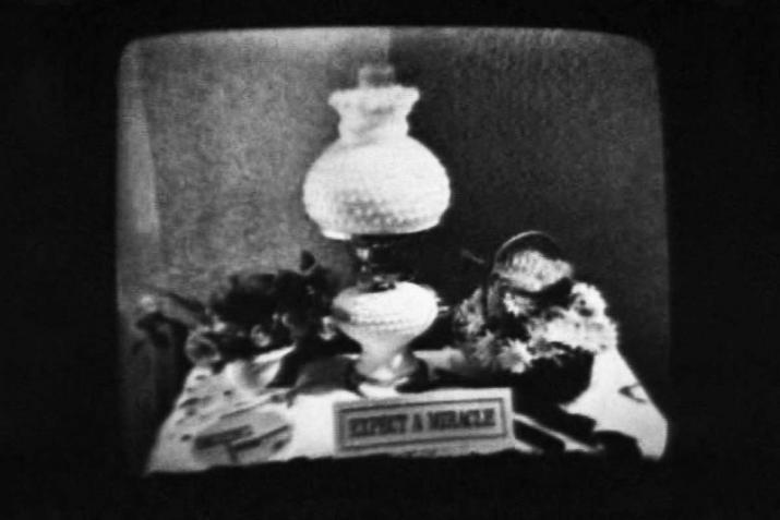 a black and white image of a lamp on a messy desk and a small sign that reads "expect a miracle"