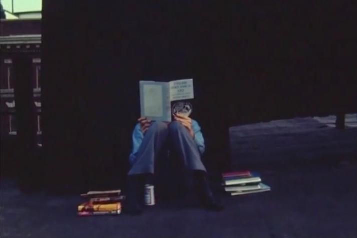 a person sitting on the ground reading with their back against a dark wall and a pile of books sitting on either side of them