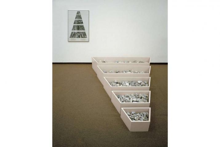 Five low bins full of rocks arranged on the floor from largest to smallest. On the wall is a framed collage of a map that shows the same shapes.