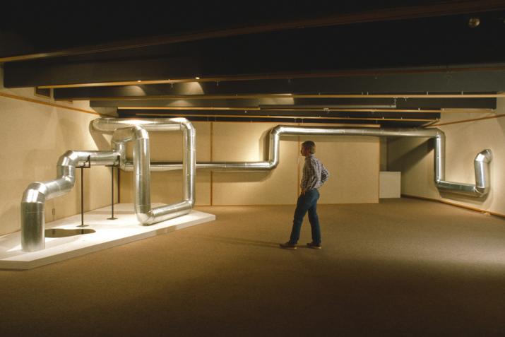 a person stands next to a sculpture made of metal pipe that zig zags through the air and along the wall in a basement room