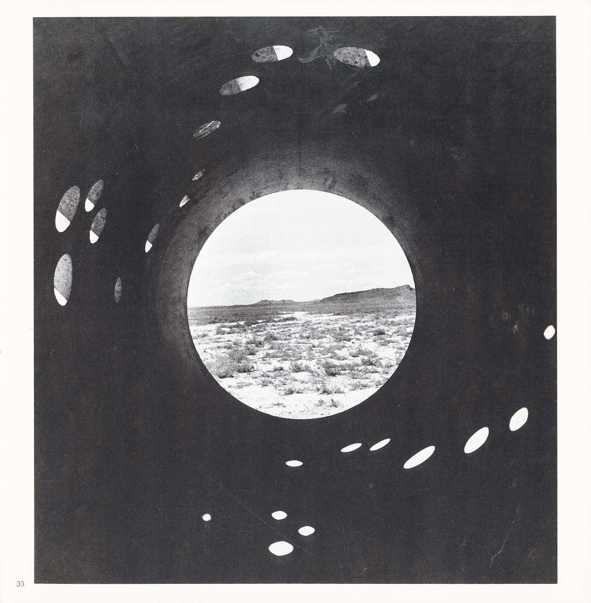 View inside a circular concrete tunnel looking out through a circular opening at the end onto the desert and mountains.  Small circular holes in the top and sides of the tunnel cast light onto the bottom of the tunnel.