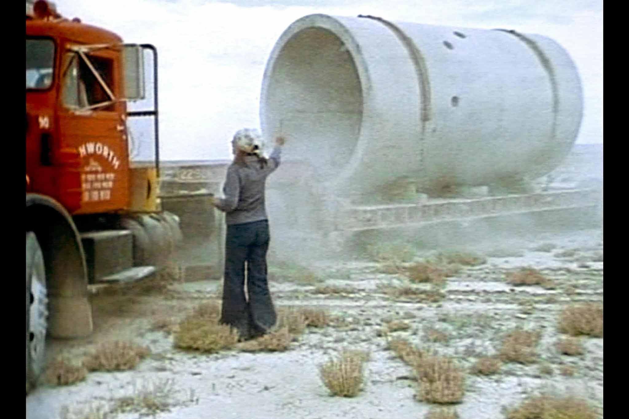 A film still from the construction of Holt's Sun Tunnels from the film, Sun Tunnels showing Holt directing a truck driver who is delivering one of the concrete tunnels.