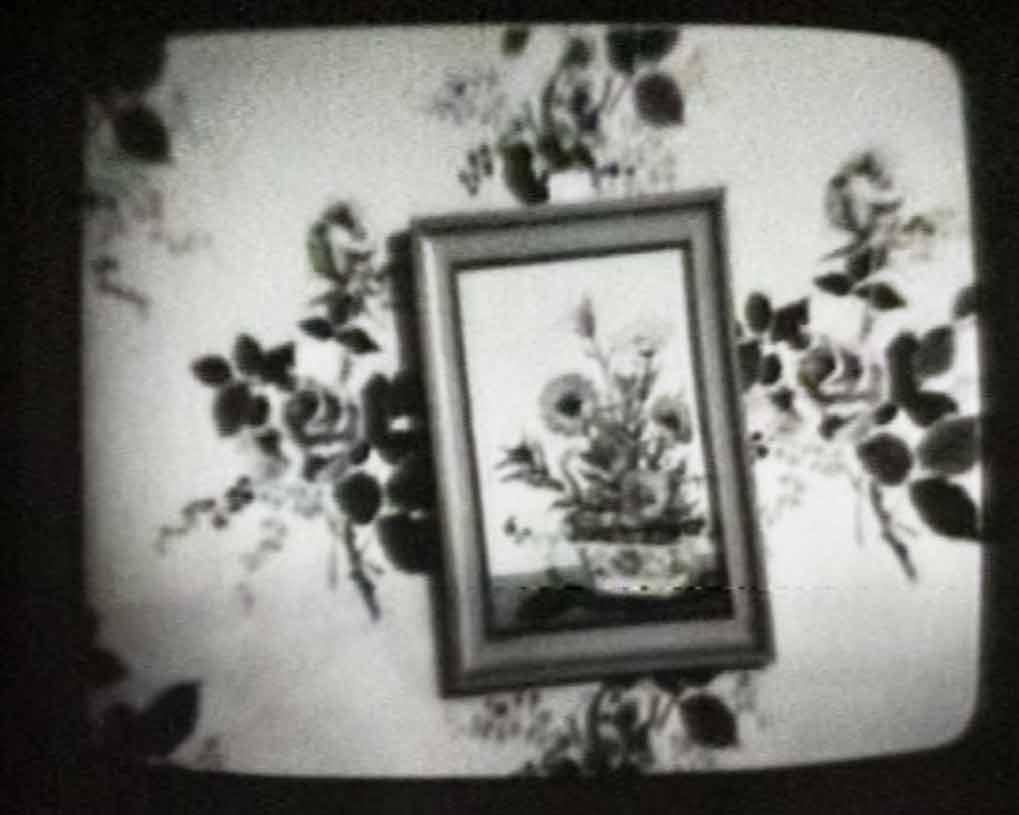 black and white image of a painting of flowers hanging crooked on a floral wallpaper