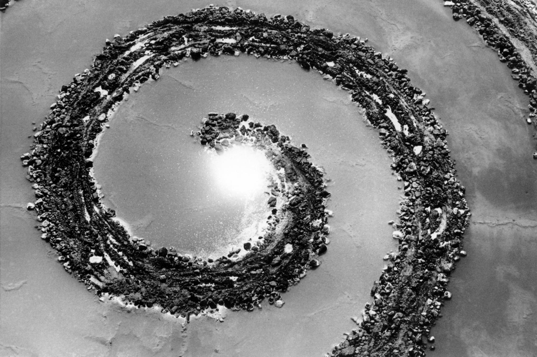 aerial black and white image of a spiral of rocks and earth in water. Sun reflected in water.