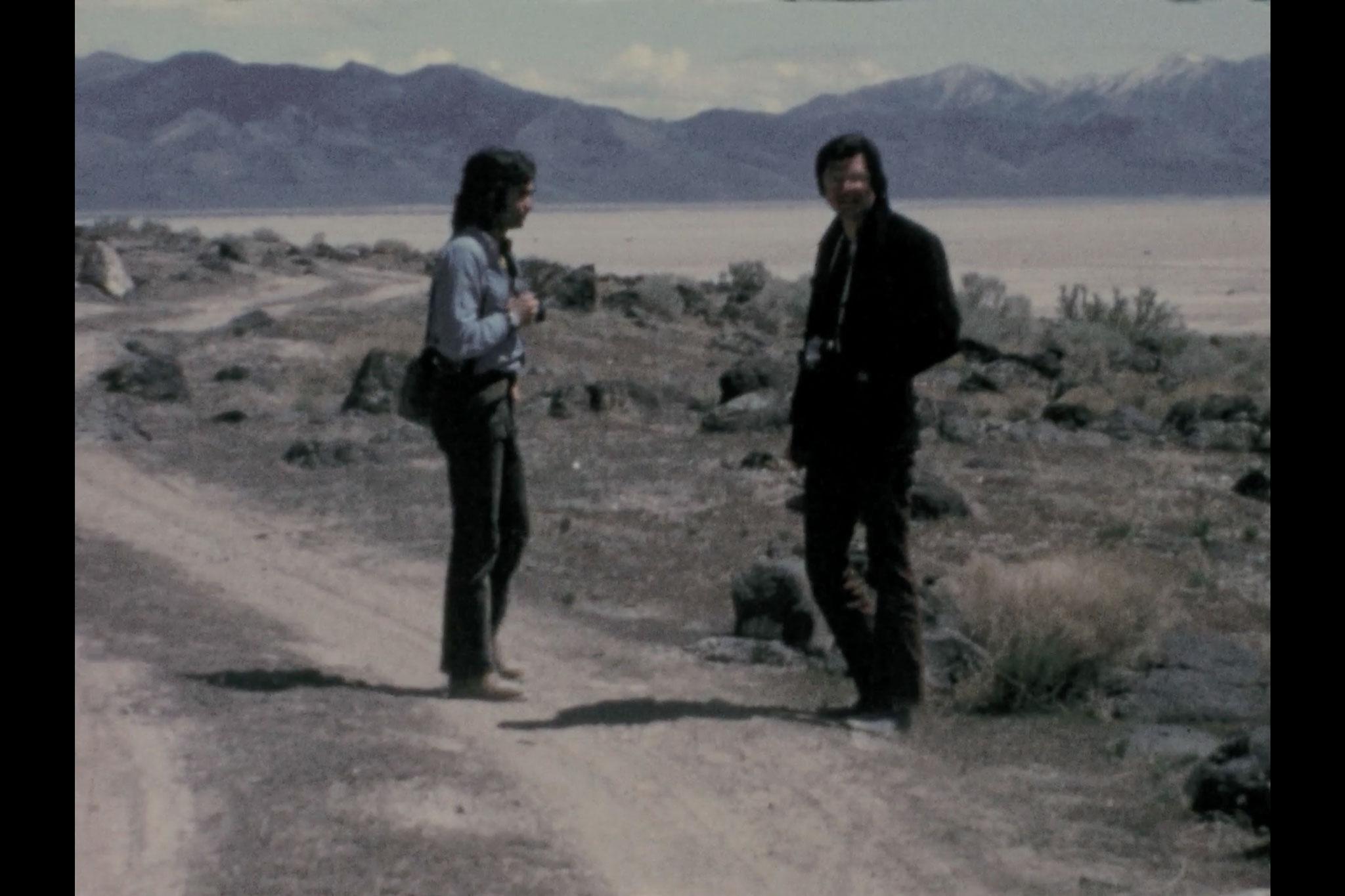 Two figures standing on a dirt road with mountains in the background