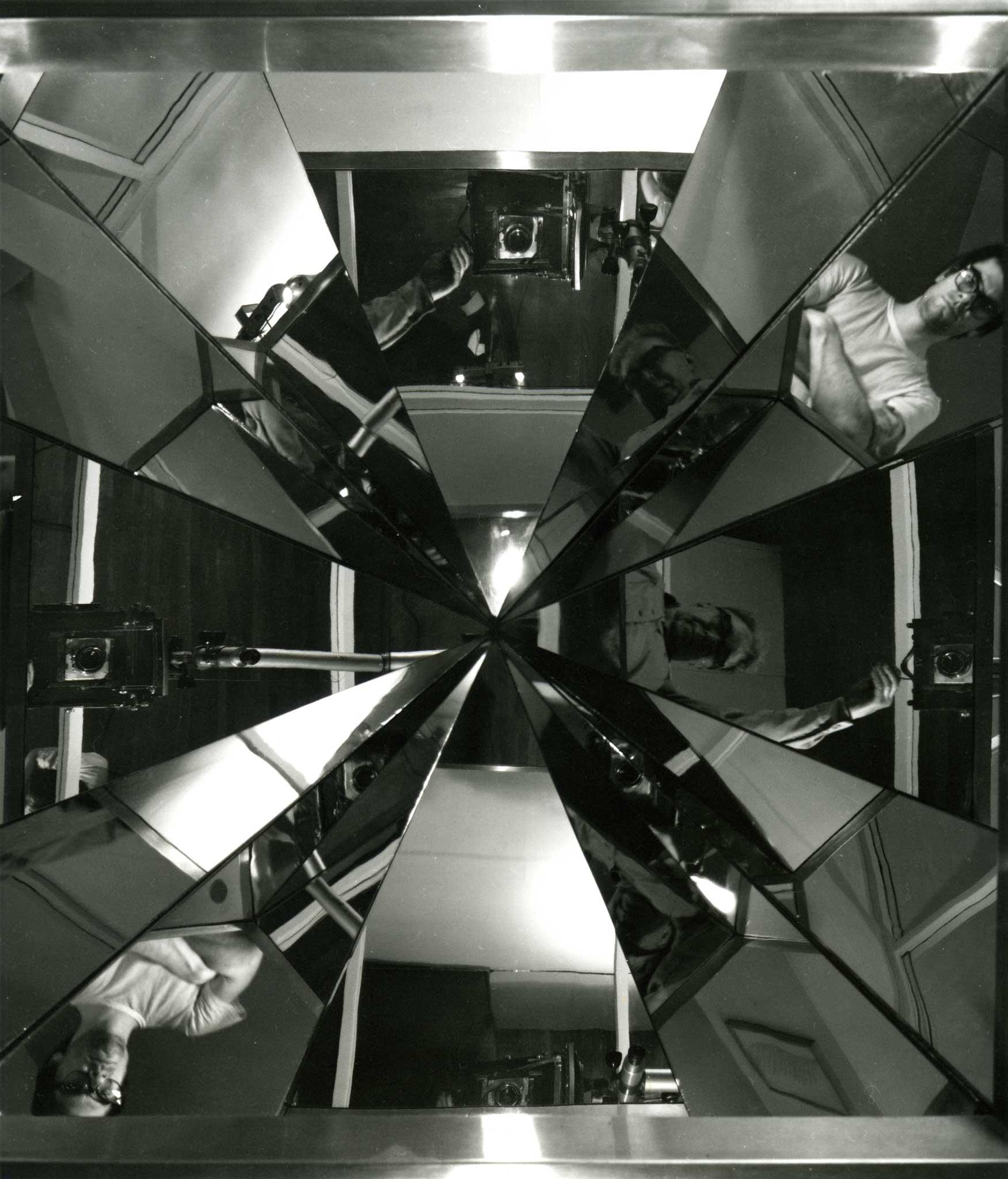 black and white image of angular reflections converging at a center point.  Reflections are of of two men standing looking into the mirrors