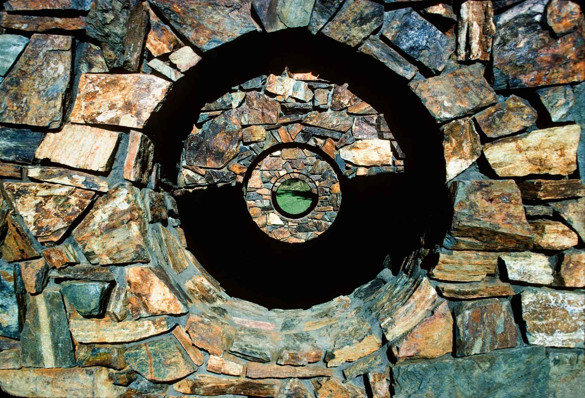 Concentric circular openings in stone walls in the center of the images. Brown stone and dark shadow and grass filling the centermost circle.