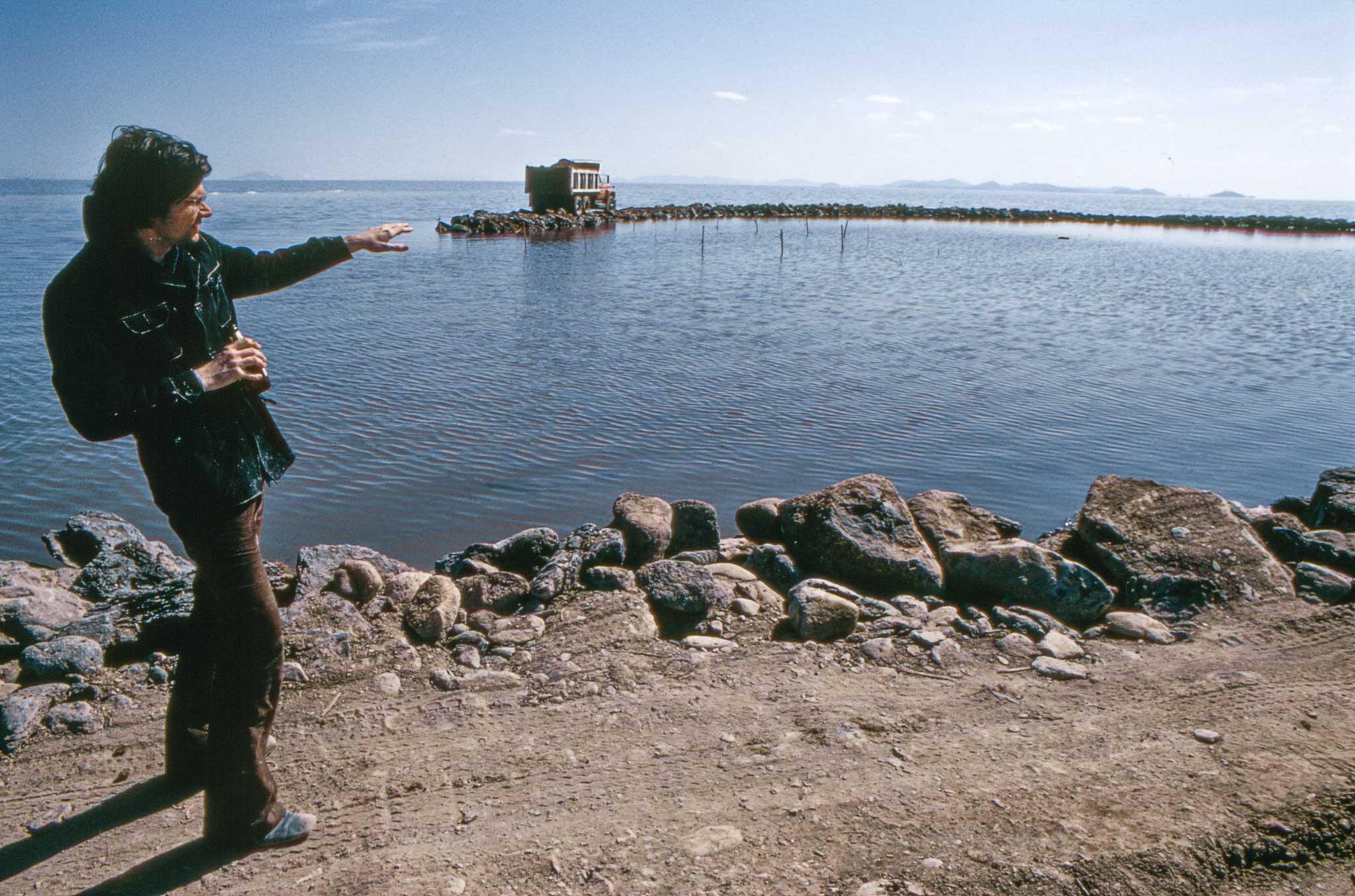 Man standing on the left side of the frame gesturing toward a jetty of land and a truck on the water