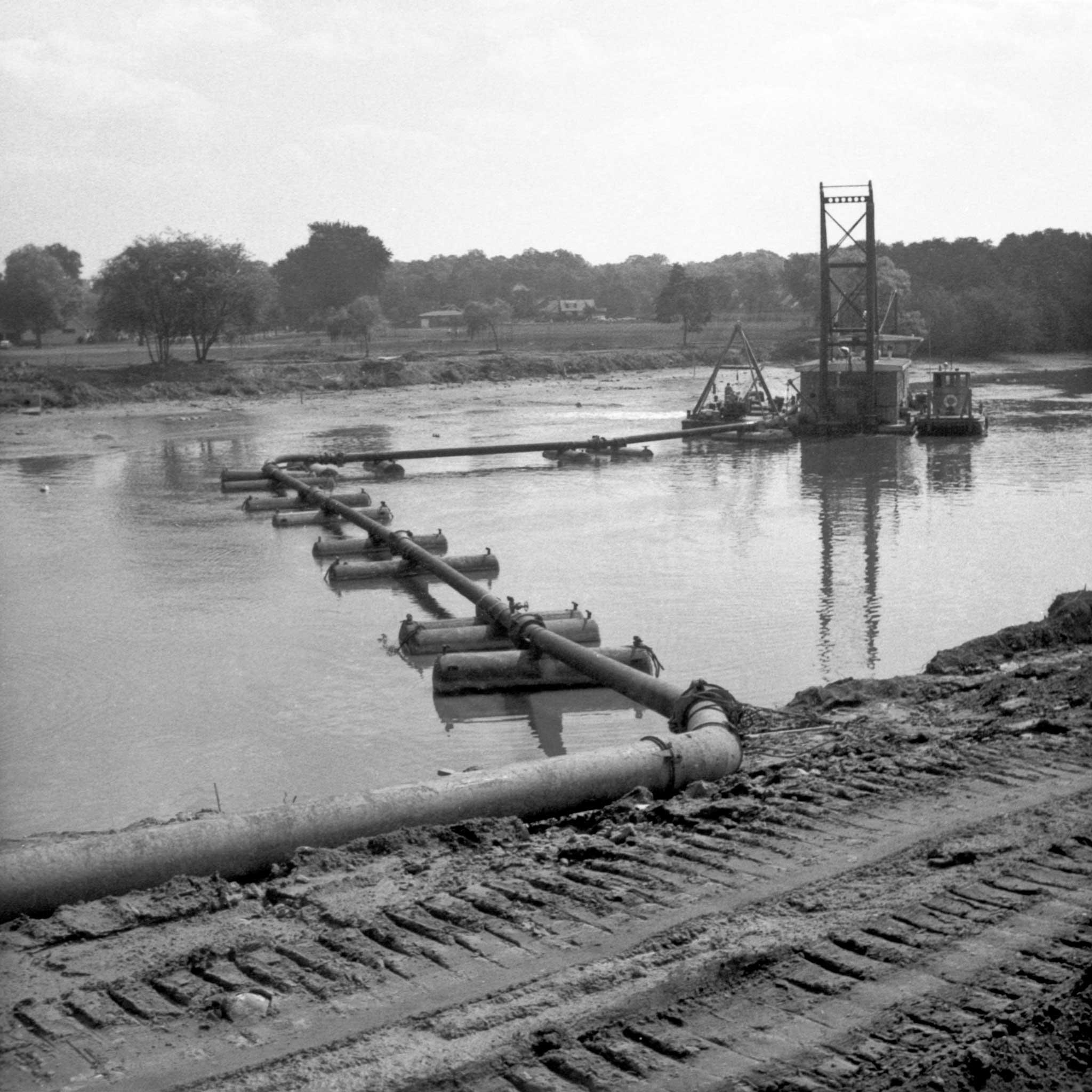 black and white image of pontoons and a pumping derrick in a body of water with trees in the background and railroad in the foreground