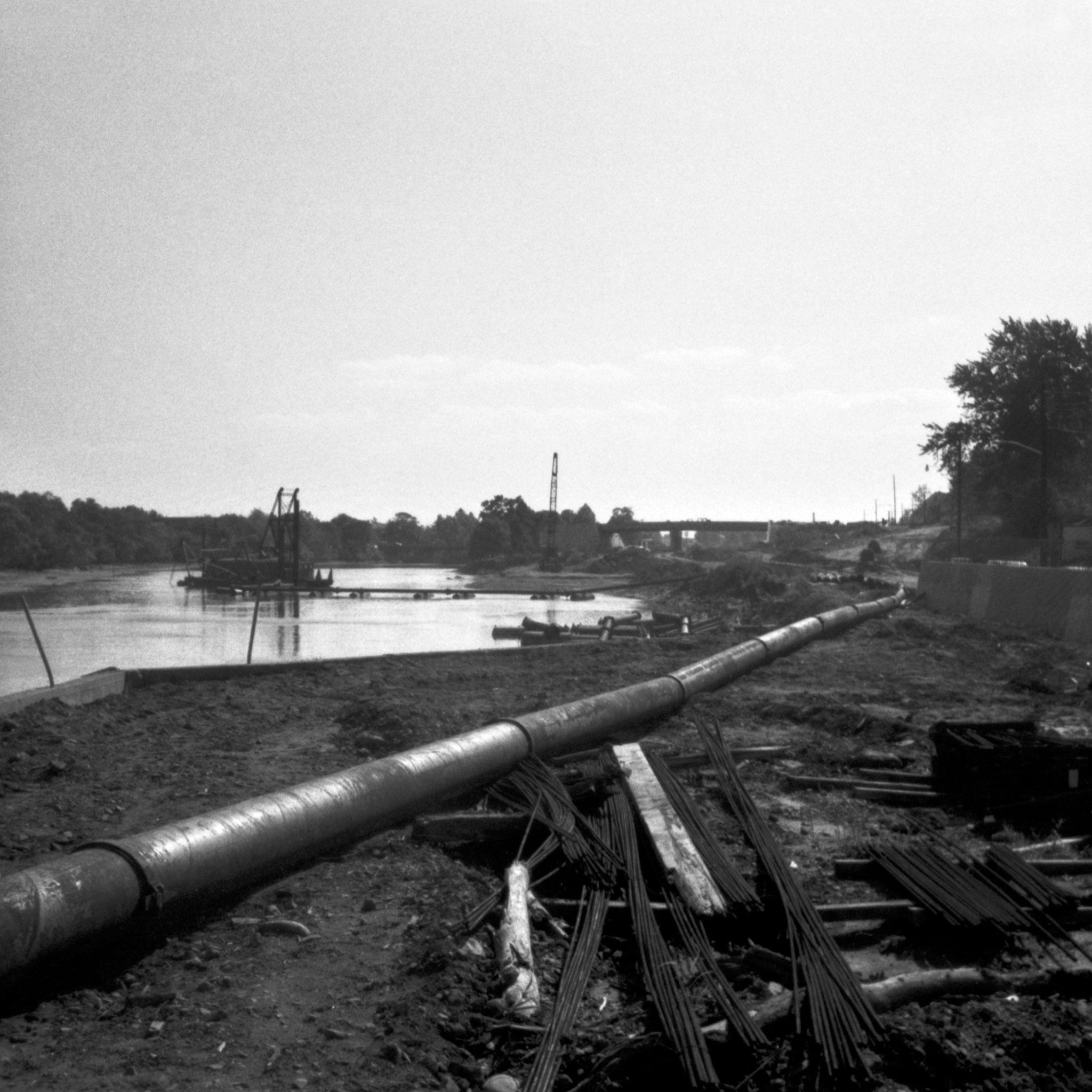 black and white image of a large metal pipe next to a body of water and landscape in the background