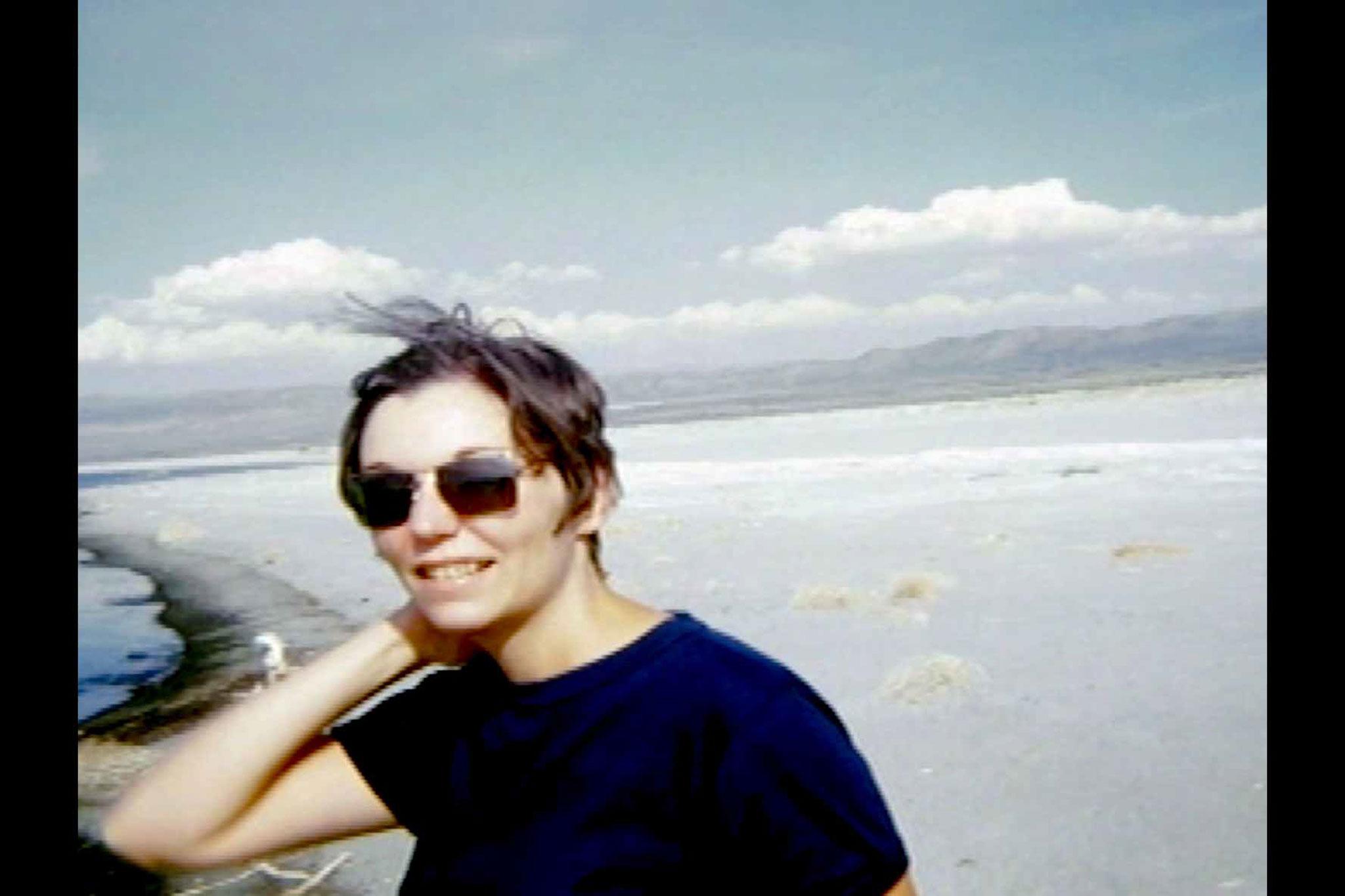 A young woman wearing sunglasses looking at the camera, with blue sky and desert behind her.