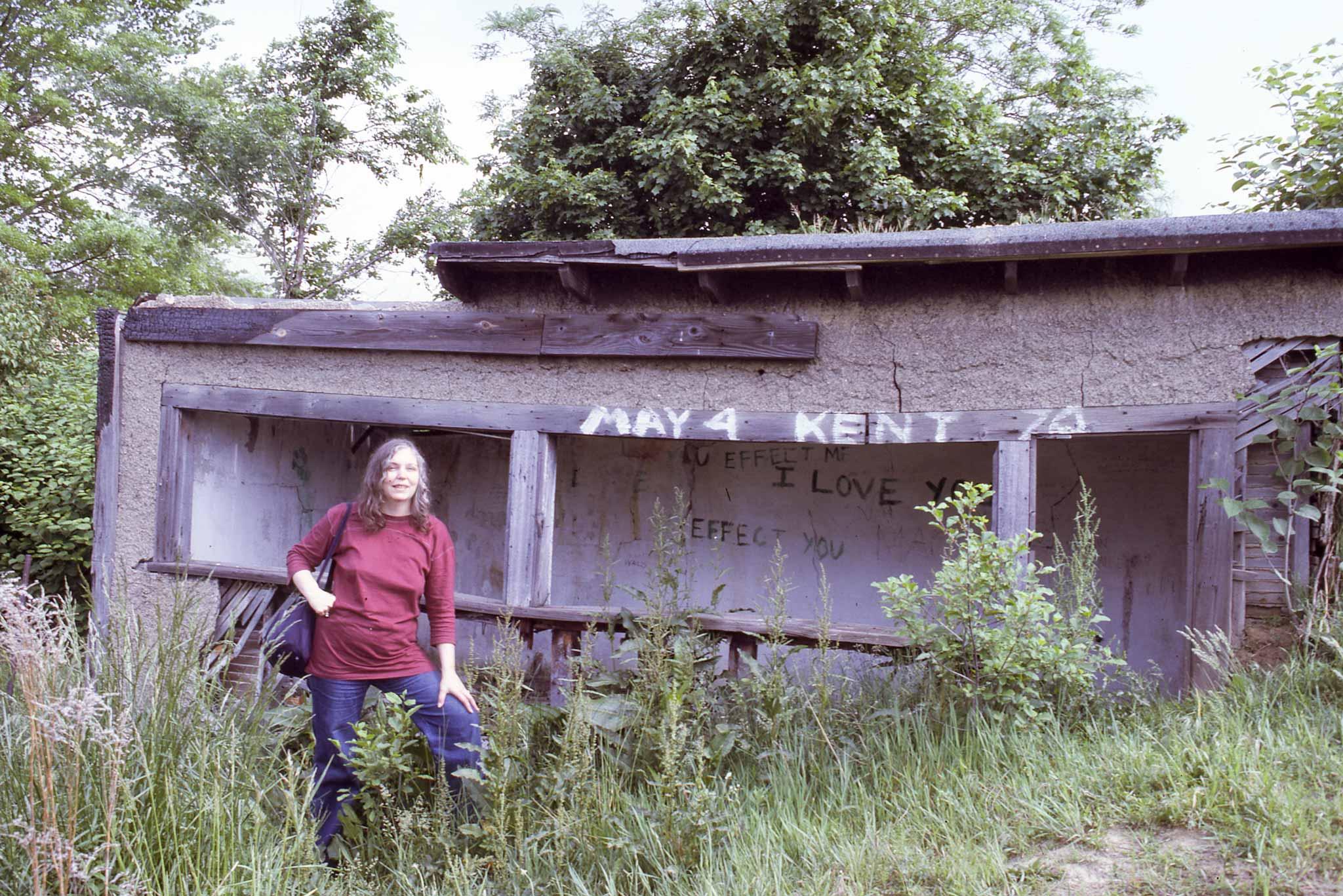 A woman stands next to a dilapidated woodshed with earth piled on the right wall and roof has been taken over by the surrounding plants