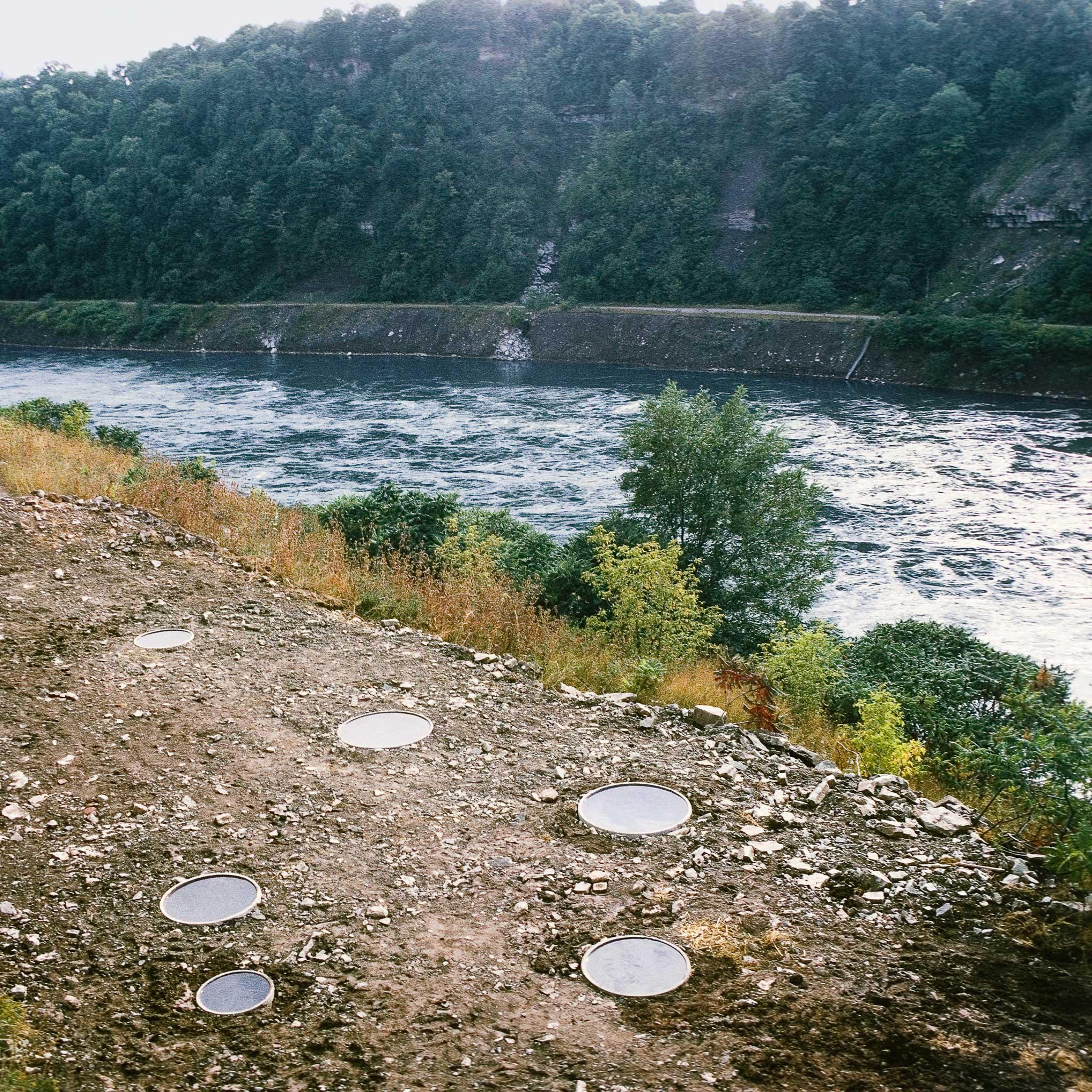 A series of small circular pools in the position of the constellation Hydra along a river bank. River and woods beyond in the background.