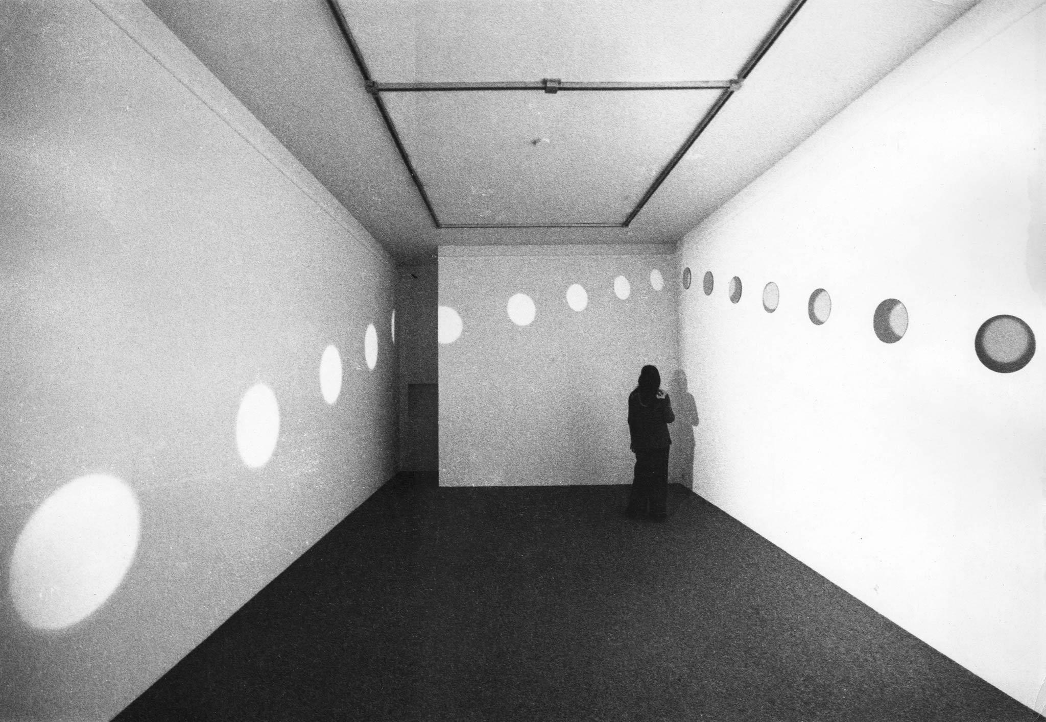 A figure standing in a white walled room. A diagonal line of mirrors in a white walled room. Light is reflecting off the mirrors, creating orbs of light on the other two walls