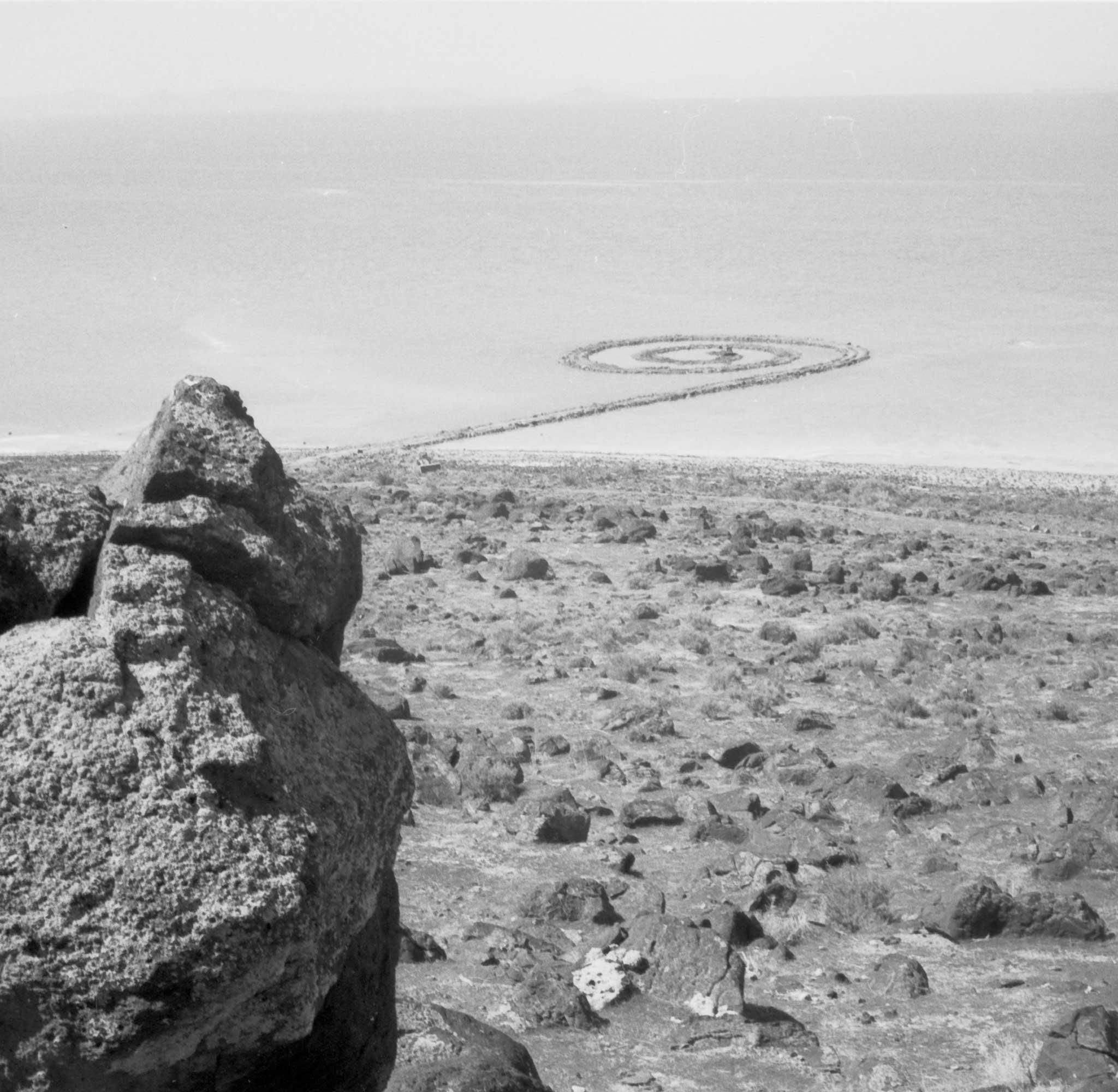 black and white image showing a spiraling jetty of rock extending out into a lake, with the shoreline and rocks in the foreground