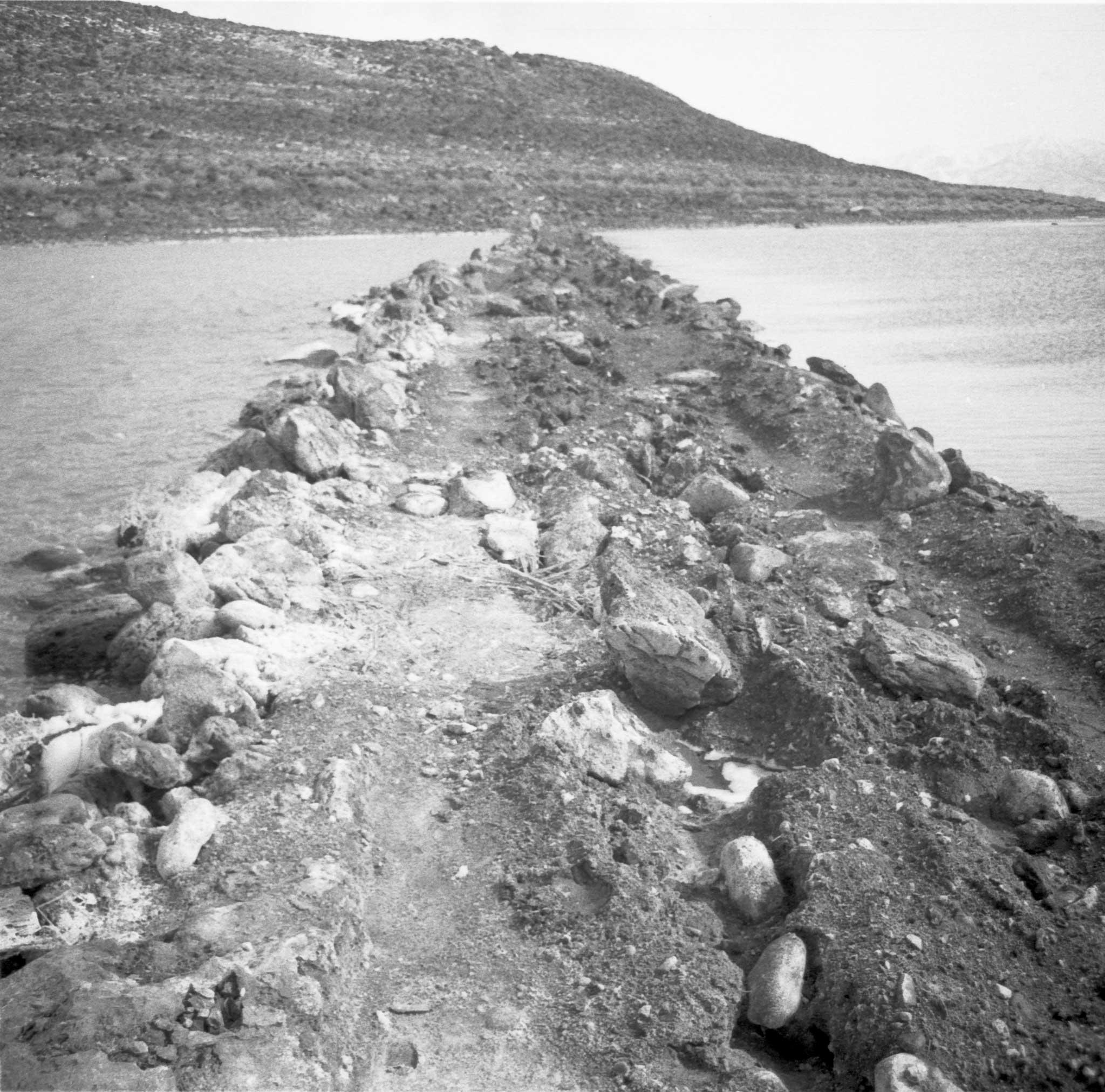 Black and white, a rock jetty surrounded by water extending straight back to the hill and shoreline