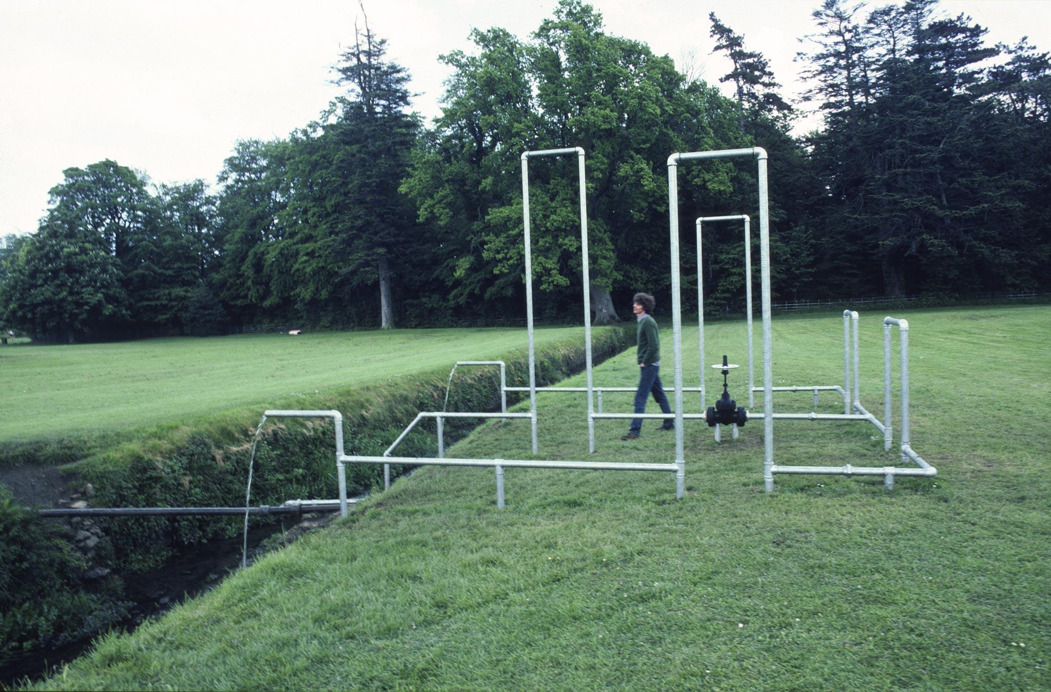 figure walking amongst galvanzied pipes organized into rectilinear structures in a field outside a large home in Ireland