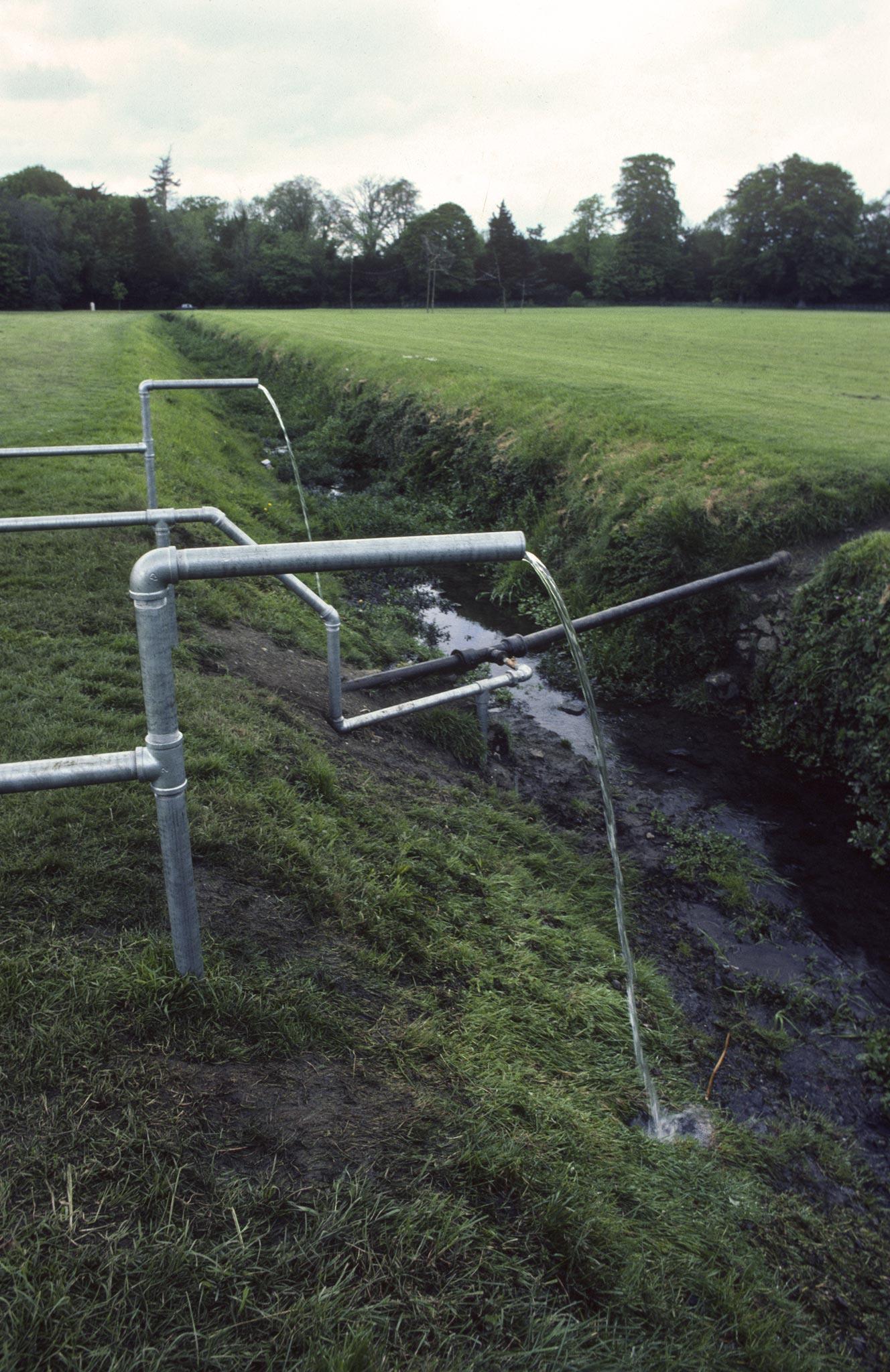 Water flowing from two galvanized pipes and dropping down into a muddy ditch