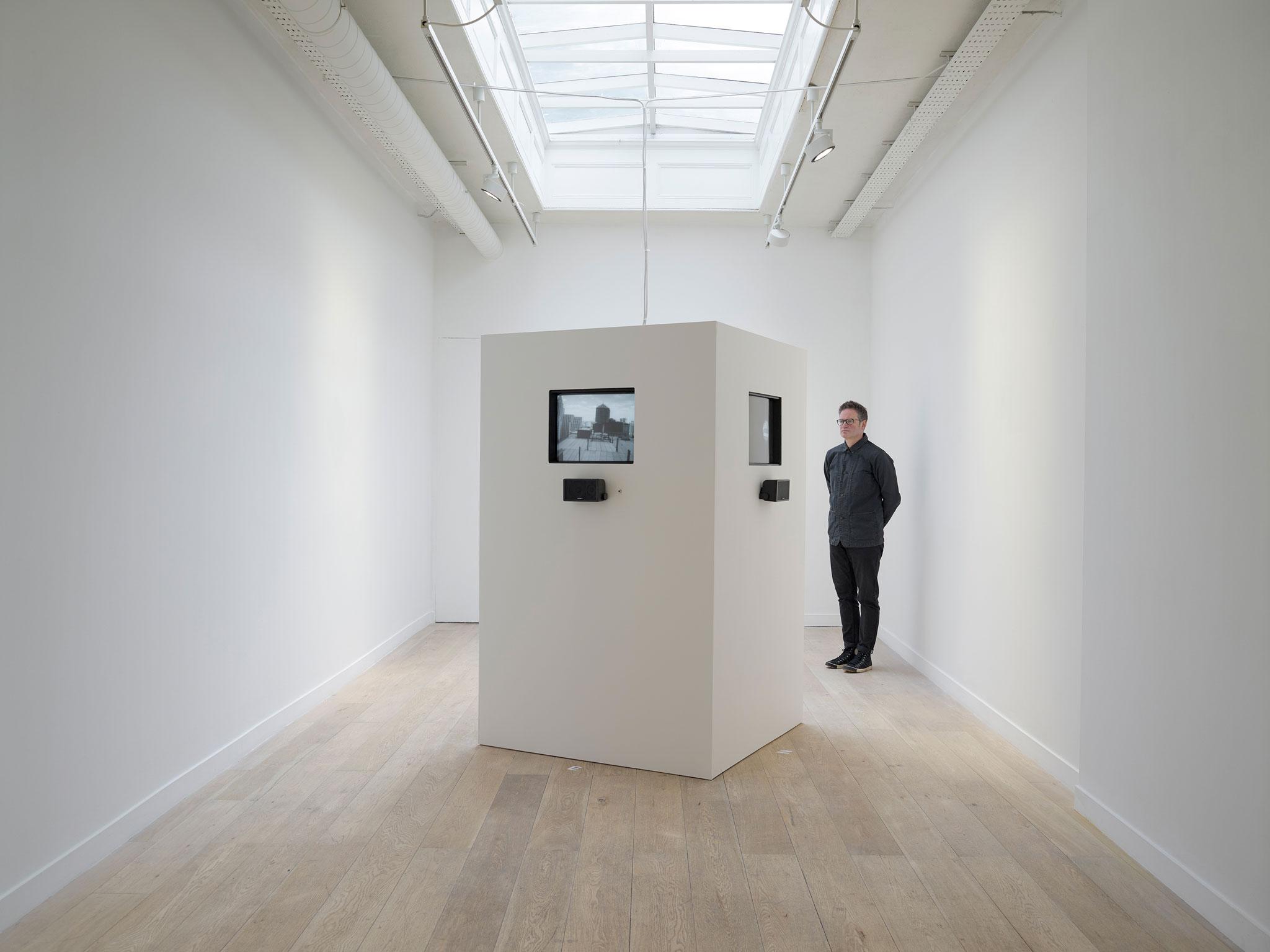 white room with a large white box in the center containing television monitors playing black and white video. One figure standing watching