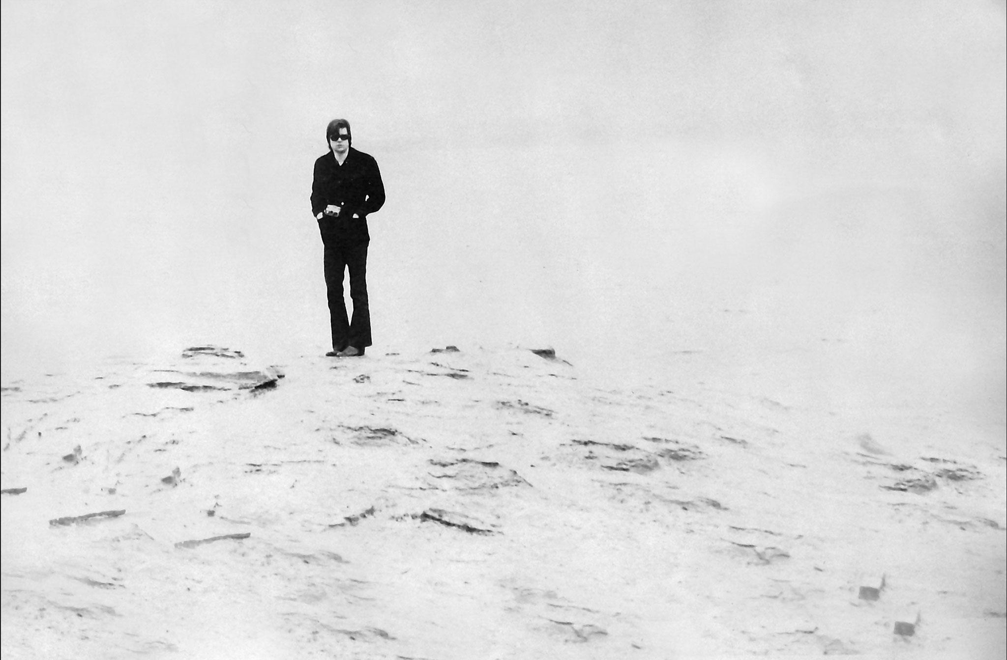black and white: a figure wearing dark clothes stands on the edge of small outcrop of land with water behind. 