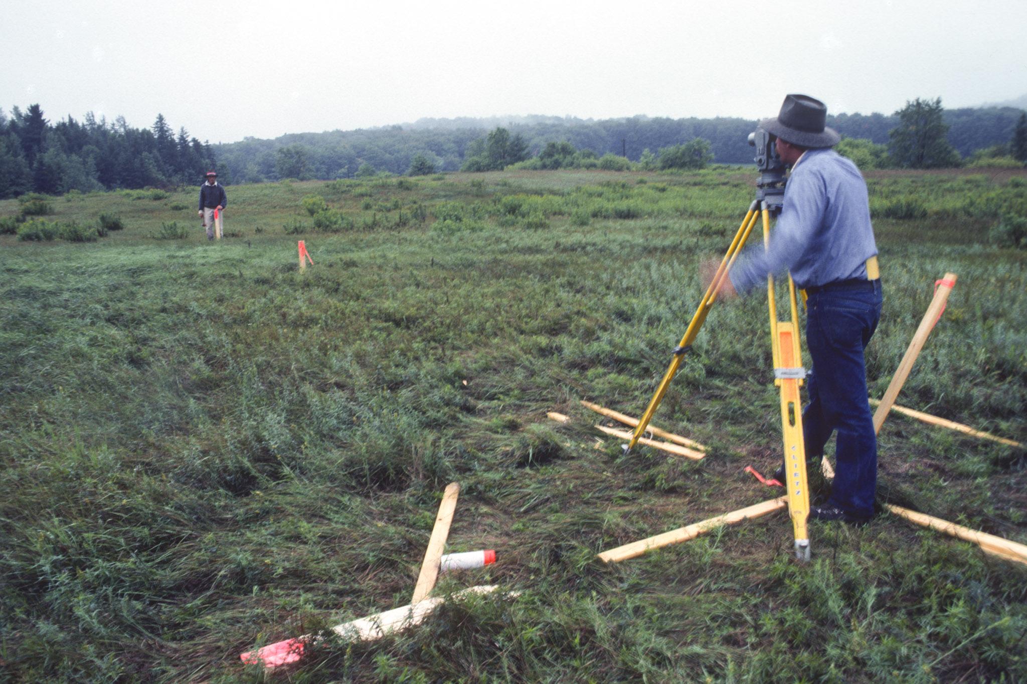 two figures stand far apart in a green field, looking through surveying equipment