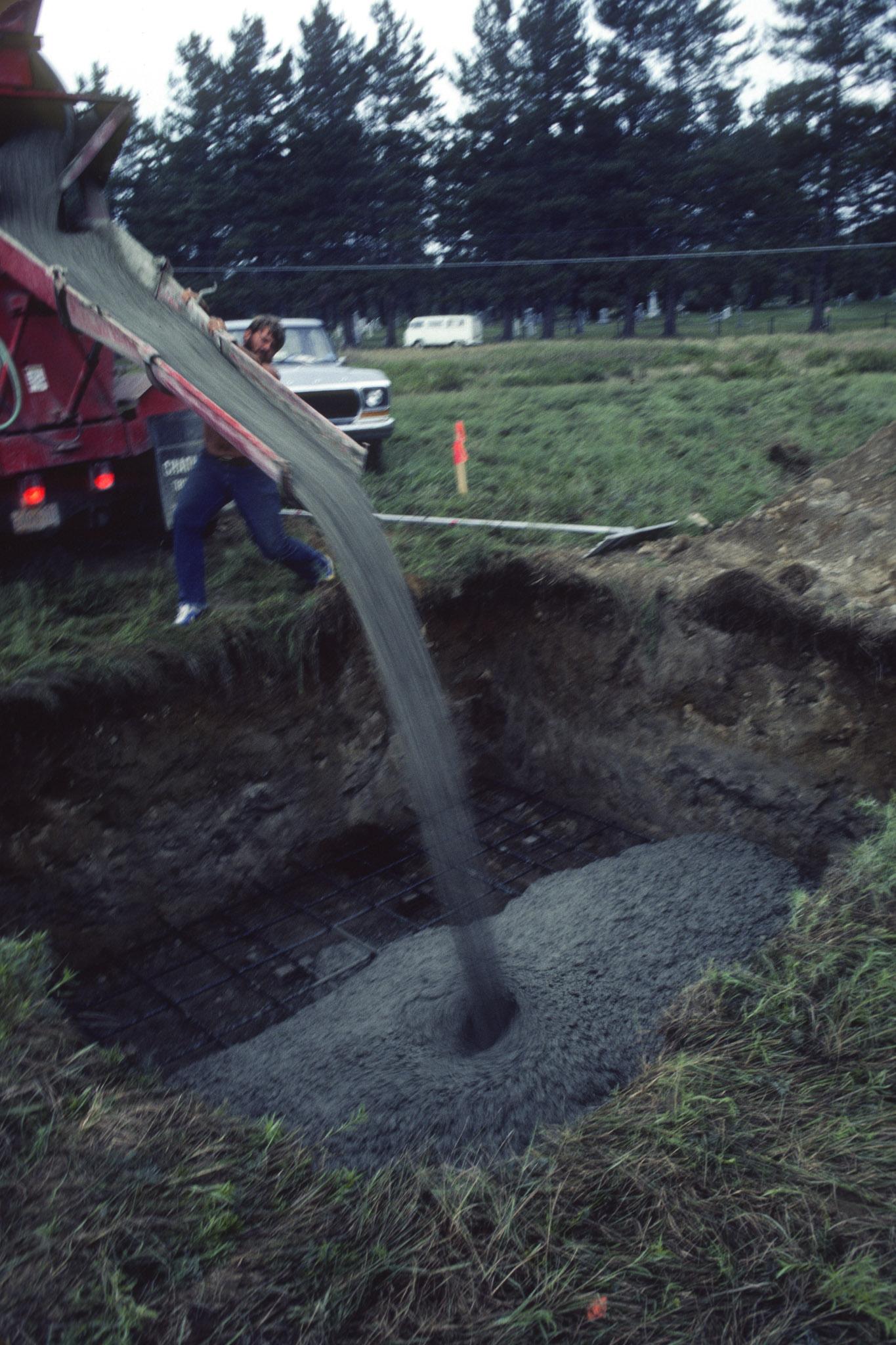 pouring concrete out of a chute into a square hole in a green grassy field