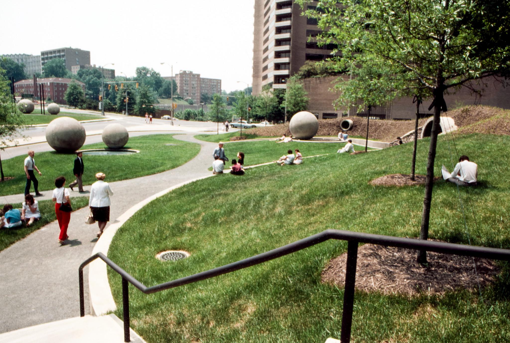 people walk and sit in a city park that features grass, small trees, and large concrete spheres