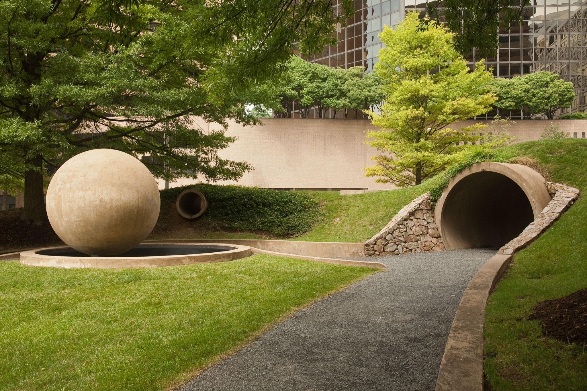 a large concrete sphere and a circular tunnel in a grassy park surrounded by trees and buildings