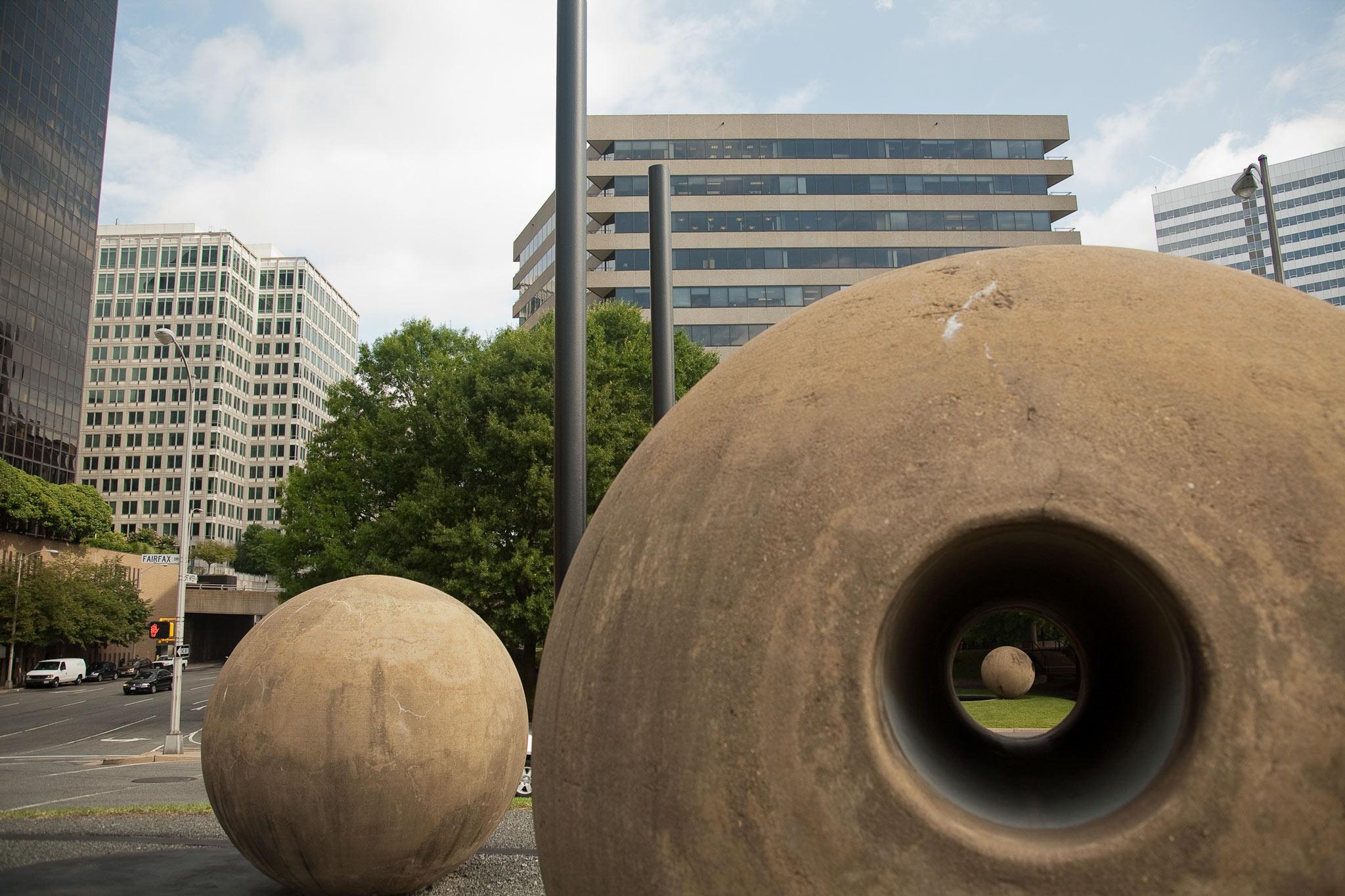 a large concrete sphere with a circular tunnel through the center that perfectly frames another concrete sphere in the distance. tall buildings in background