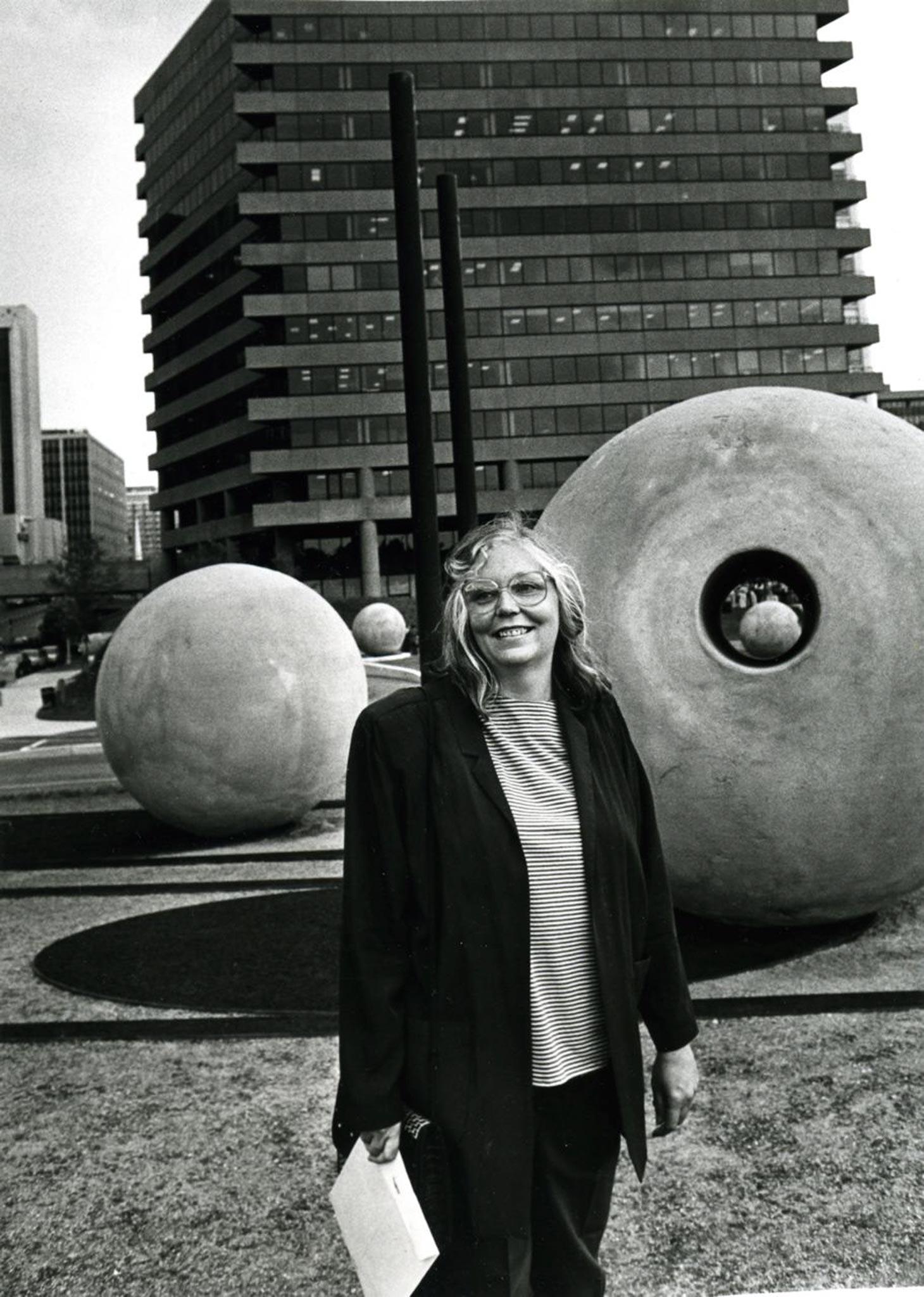 a person standing smiling at the camera. In the background are large concrete spheres and tall buildings