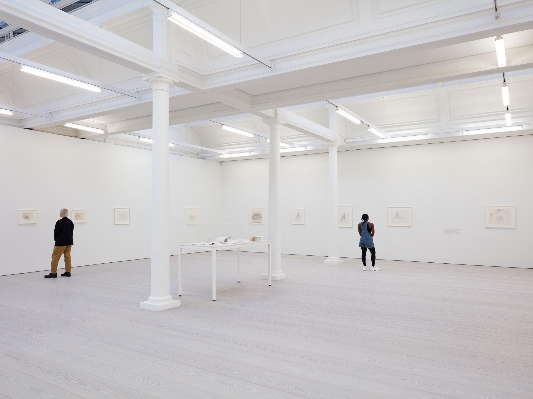 two people standing in a large white gallery space with framed drawings on the wall