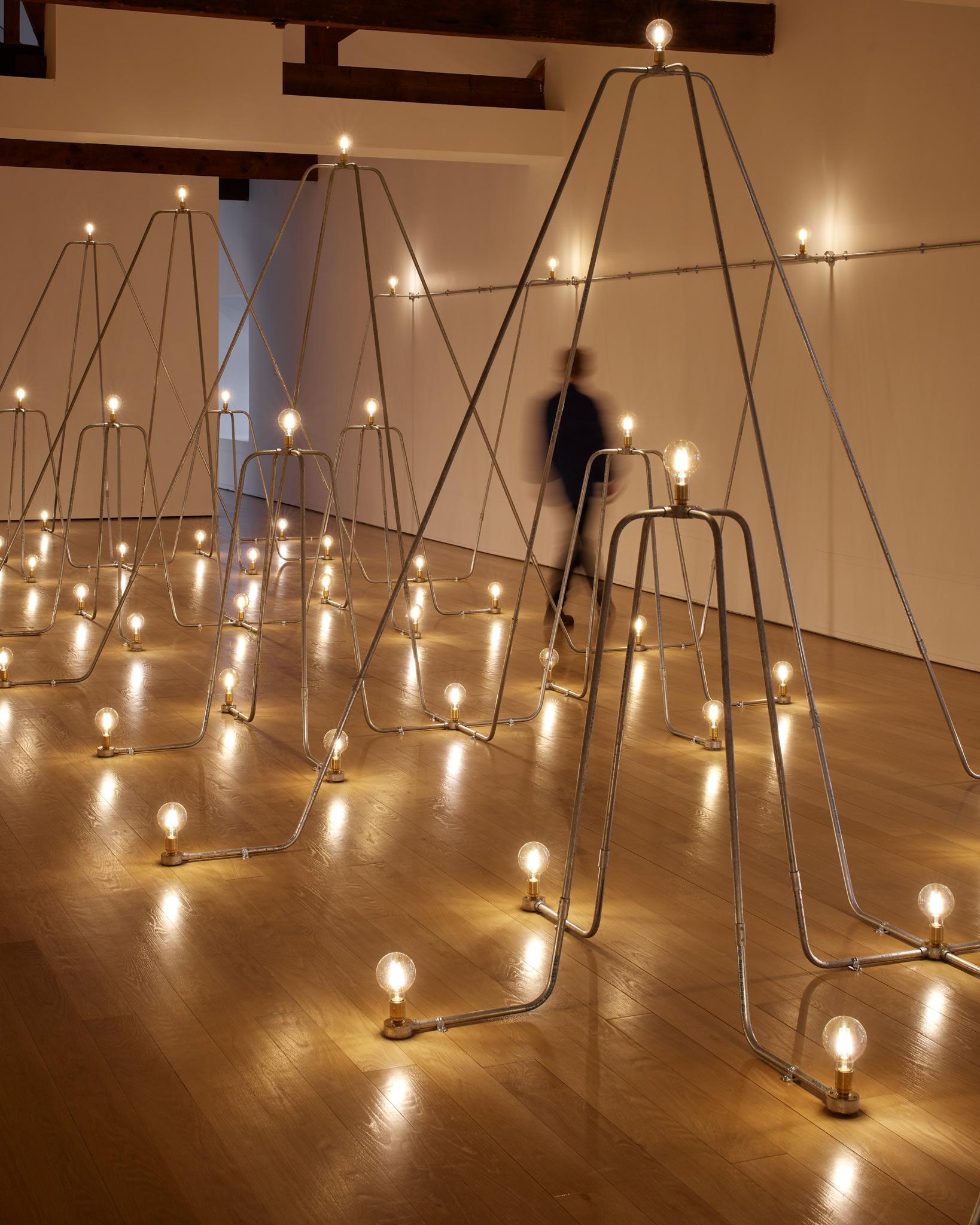 gallery space filled with arches of conduit with glowing yellow lightbulbs 