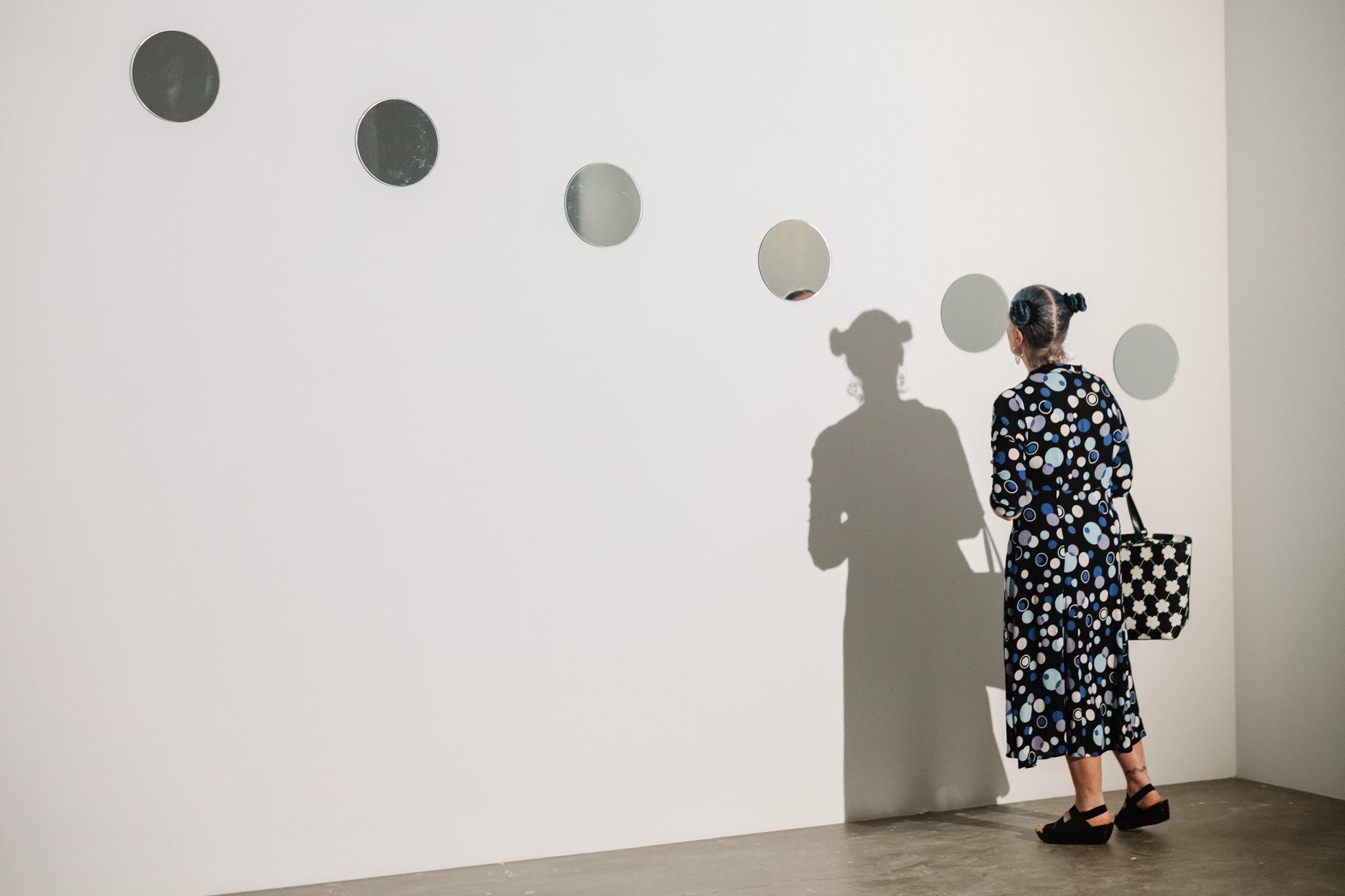 person in a gallery space looking at a diagonal line of small circular mirrors placed on the wall