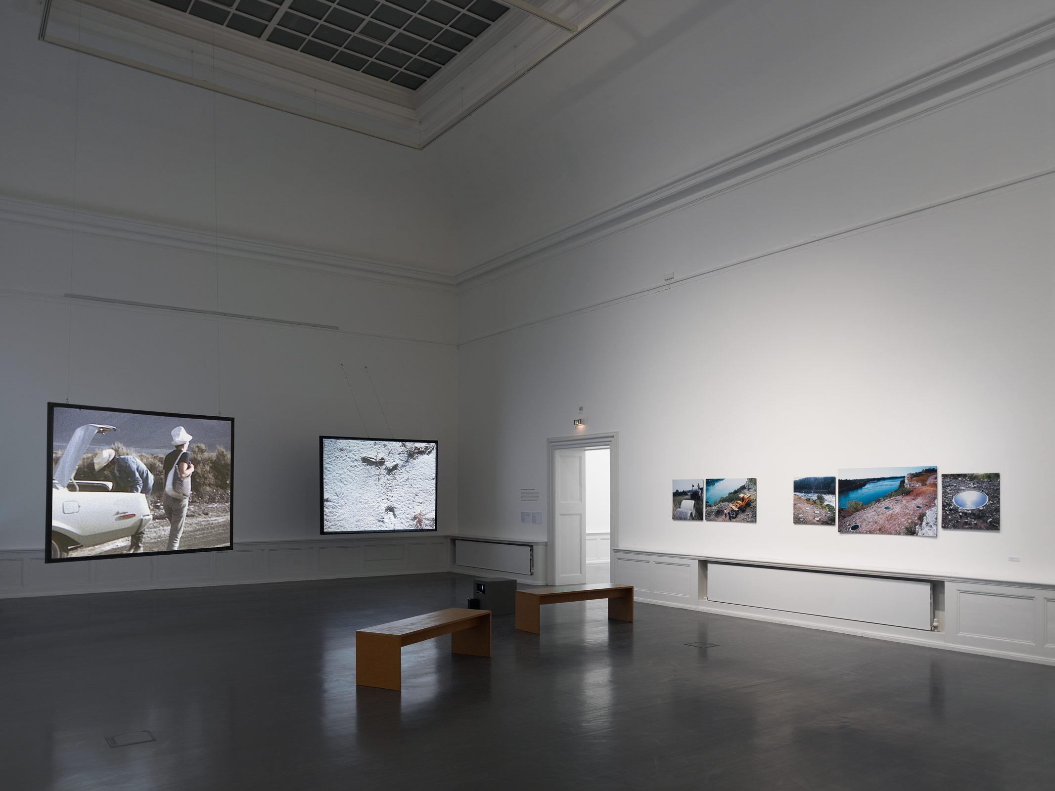 A large white walled room with benches and projected images on two hanging screens and images on the wall.