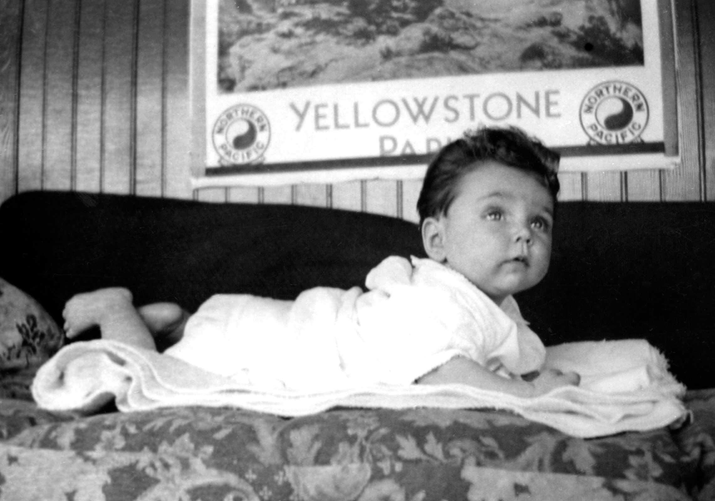 Nancy Holt as a young baby laying on a sofa with a Yellowstone Park poster in the background.