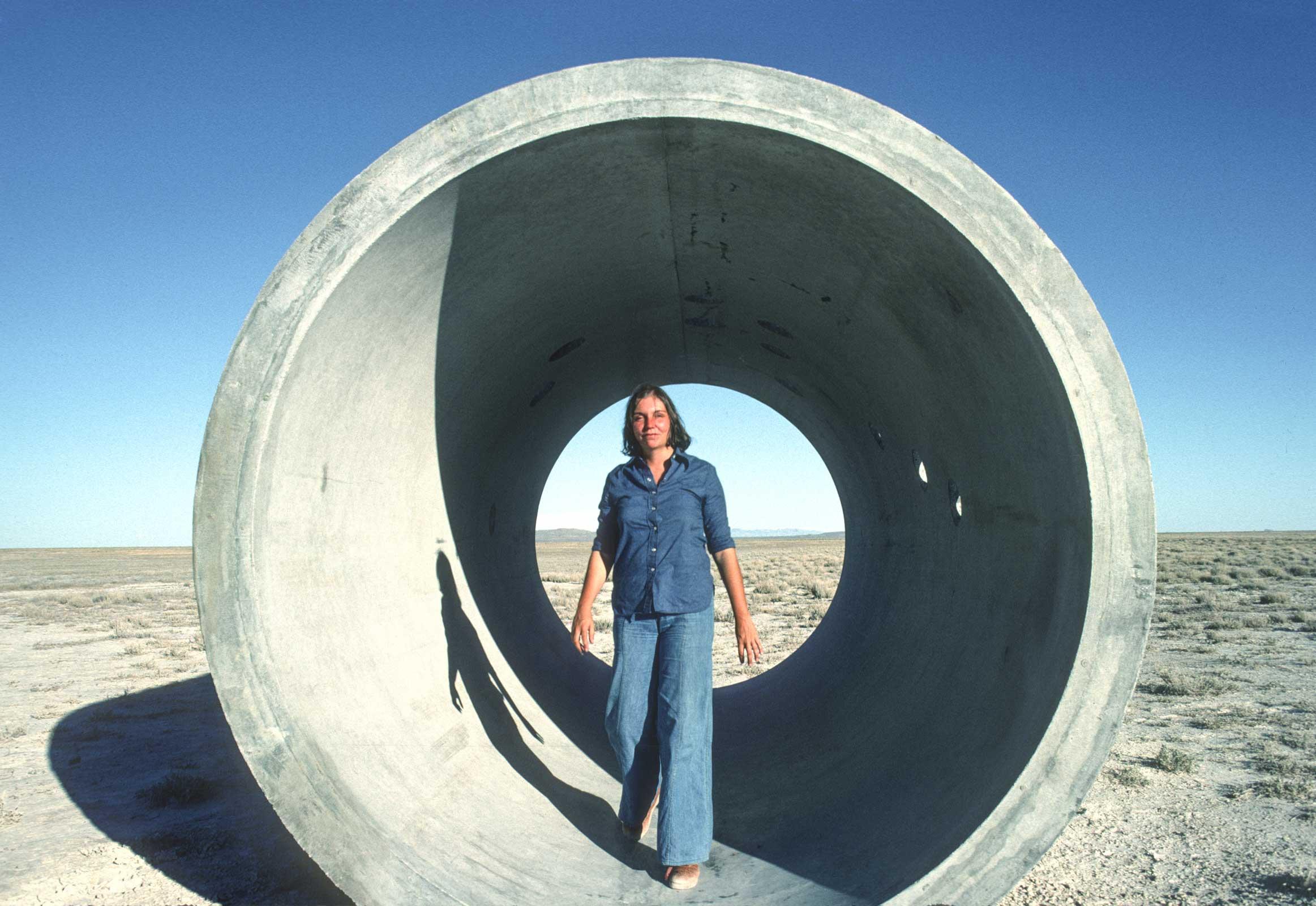 Nancy Holt standing inside one of the large tunnels that are part of Sun Tunnels.  Blue sky and sunny and Nancy is wearing a blue top and blue jeans.