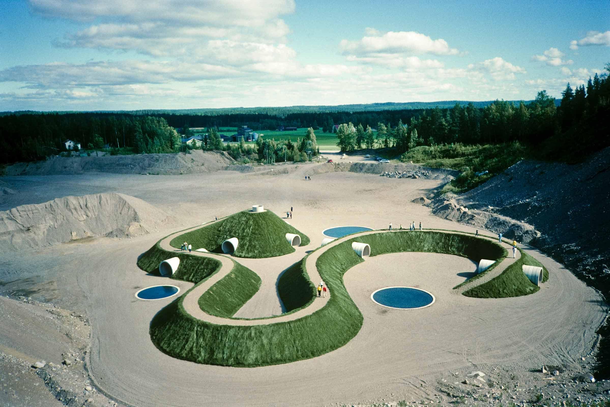 large earthwork sculpture made using earth, concrete, water, and grass with people moving around on top of it.