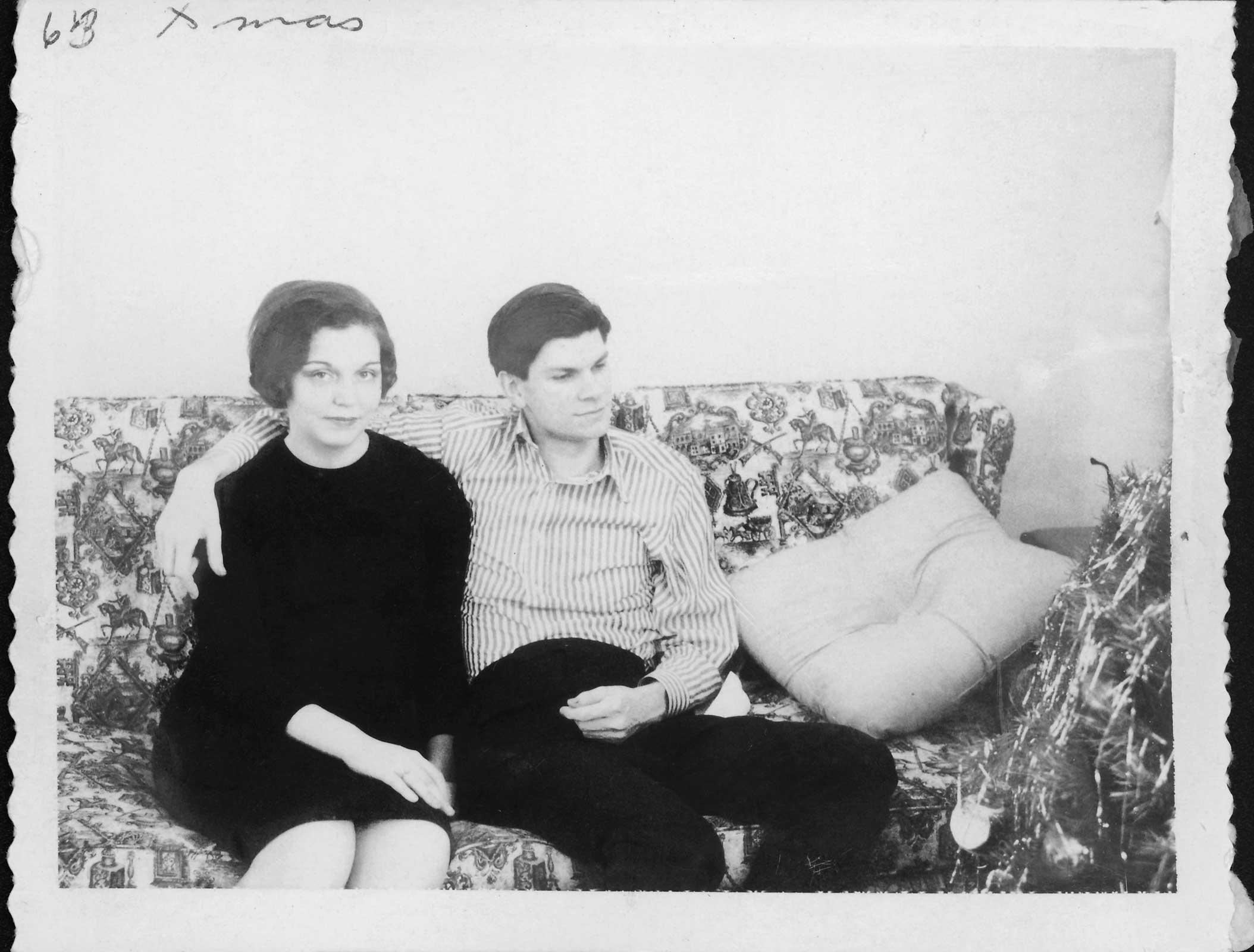 black and white photograph of a man and woman sitting on floral pattern sofa. Man has his arm around the woman and she is looking at the camera.