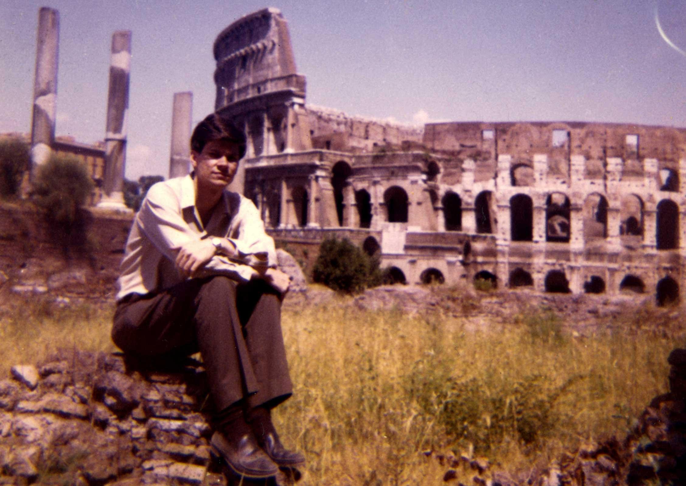 a young man seated on a rock with the Colosseum in Rome in the background
